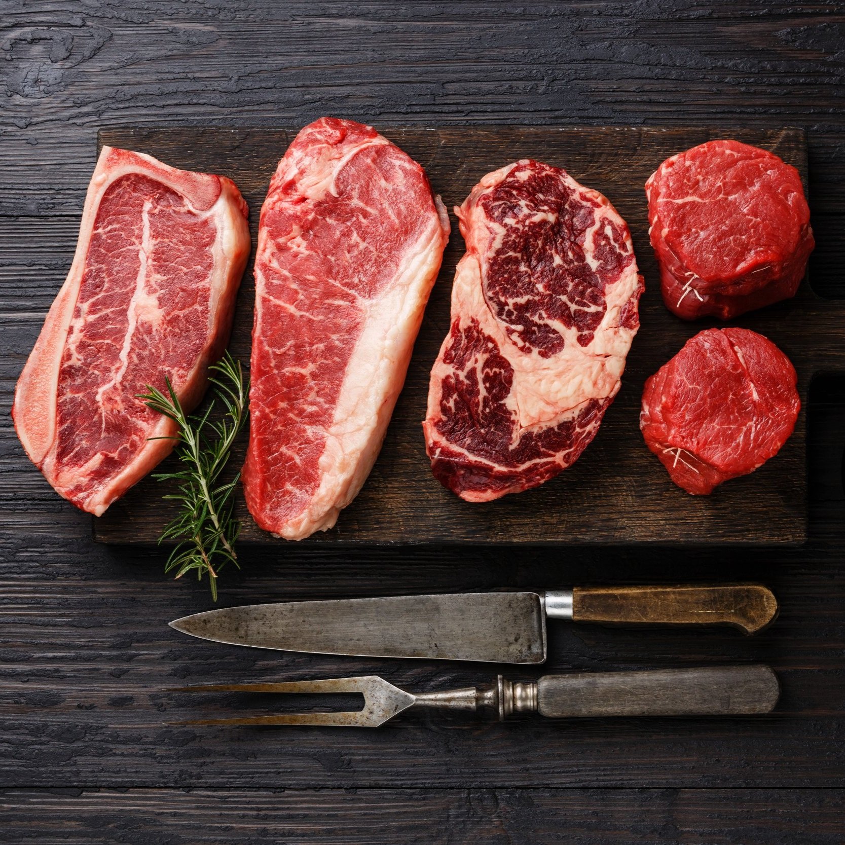 Assorted cuts of raw beef including ribeye, sirloin, and tenderloin displayed on a wooden cutting board with a sprig of rosemary and vintage meat carving tools.